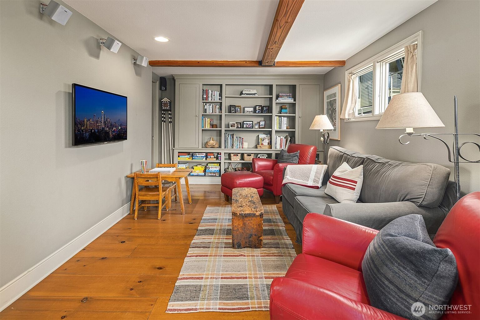 This is an inviting living room featuring hardwood floors, a built-in bookshelf, and comfortable seating including a gray sofa and red armchairs. A flat-screen TV is mounted on the wall, and a plaid rug adds warmth to the space. The room has a cozy and relaxed atmosphere, perfect for family gatherings or quiet evenings.