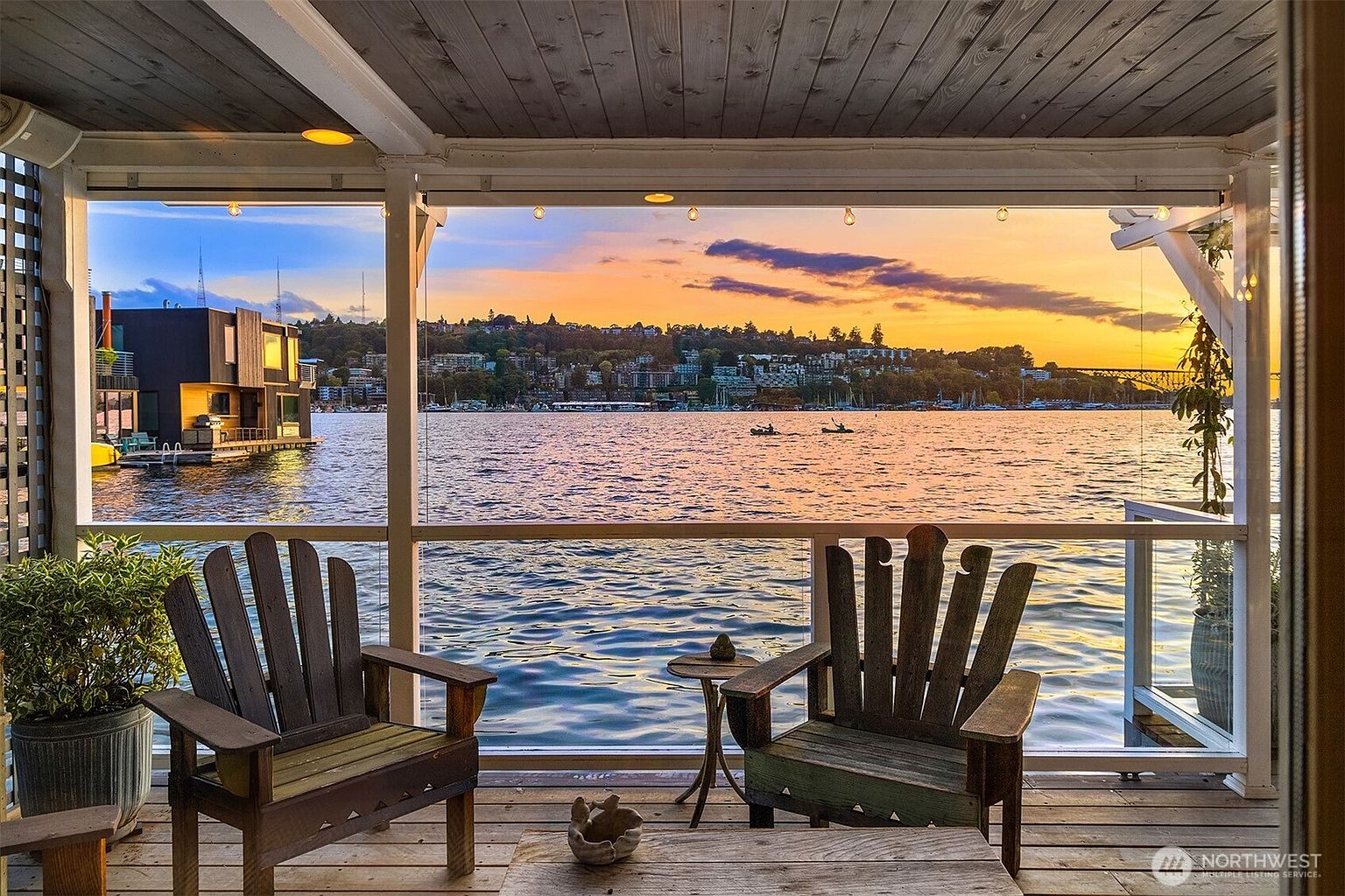 This image showcases a serene patio or deck overlooking a tranquil body of water at sunset. Two wooden Adirondack chairs invite relaxation, complemented by a small side table and a wooden coffee table. The view includes a cityscape and a bridge in the distance, creating a picturesque and inviting outdoor living space.