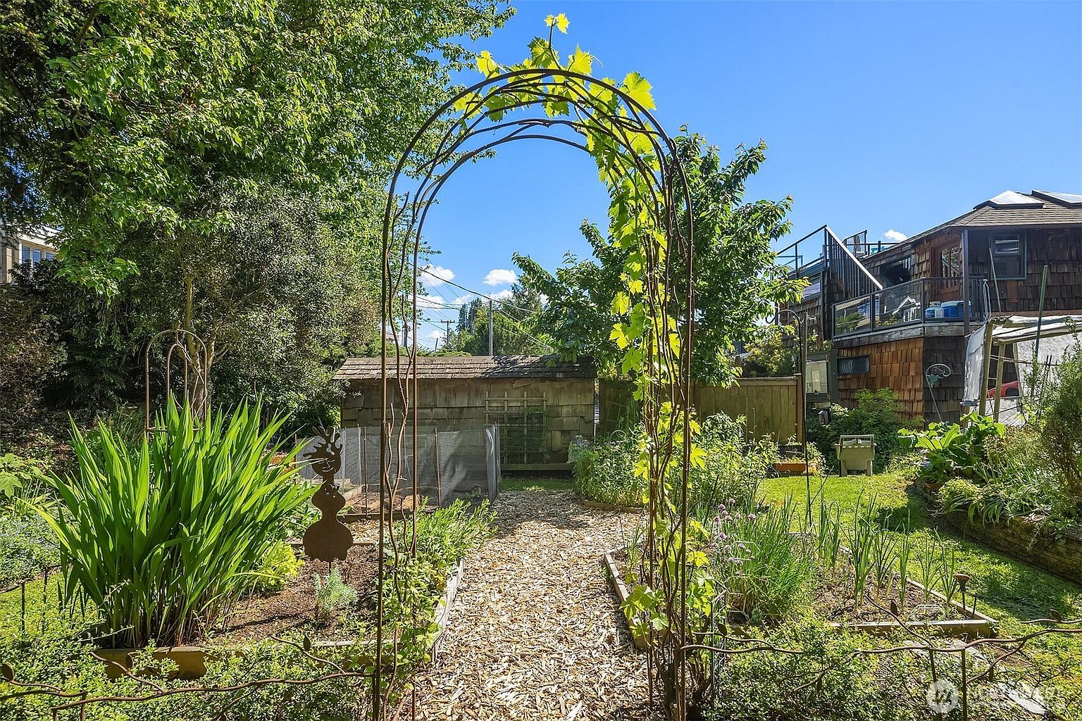 This image showcases a charming garden with a rustic aesthetic. A metal archway covered in vines serves as an entrance to a path lined with wood chips, leading towards a shed. Raised garden beds filled with various plants and greenery add to the garden's appeal, creating a tranquil and inviting outdoor space.