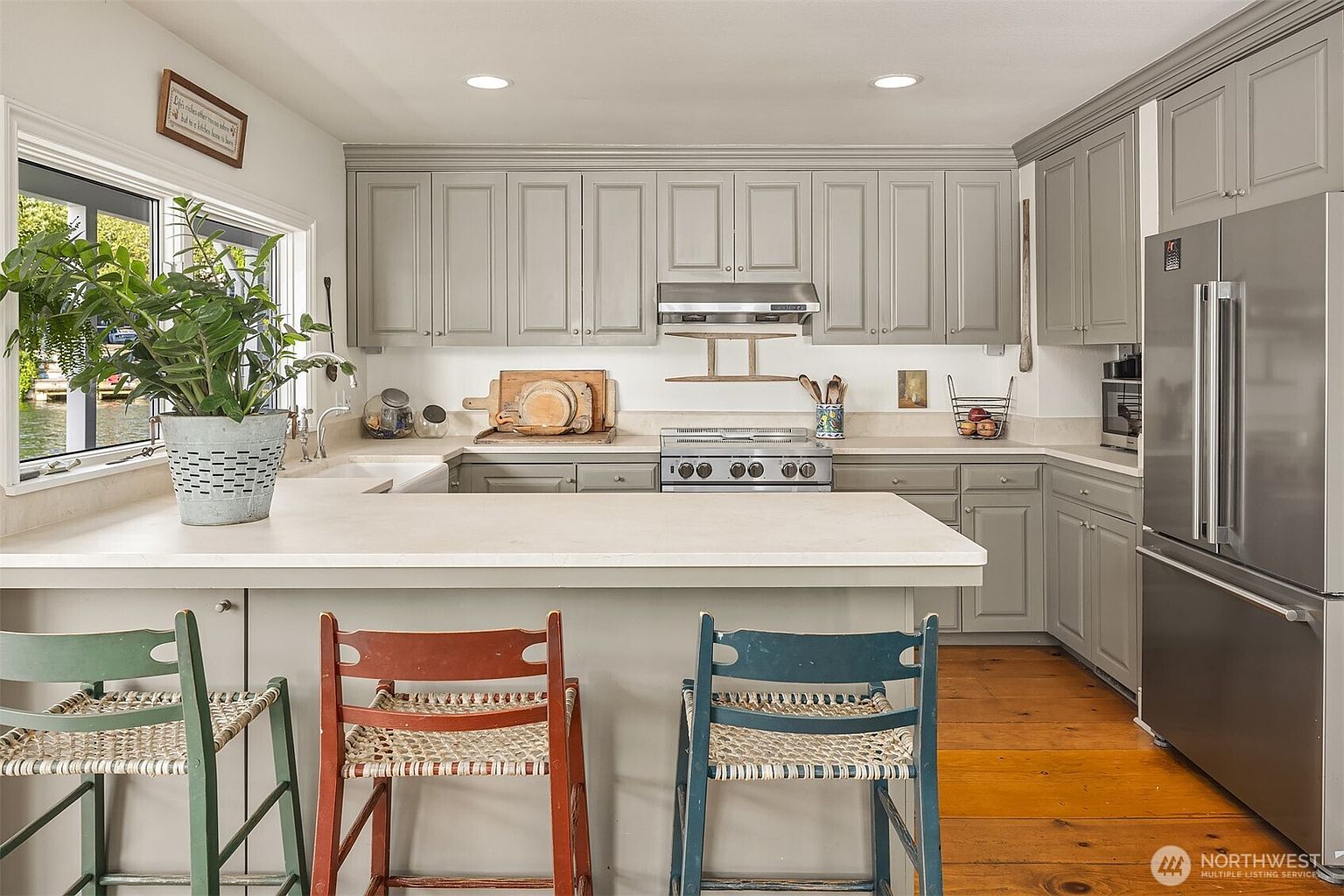 This is an interior shot of a kitchen featuring gray cabinets, stainless steel appliances, and a light-colored countertop. Three colorful chairs are positioned at the kitchen island, adding a touch of character to the space. The kitchen has a cozy and inviting feel, with natural light streaming in from a window.