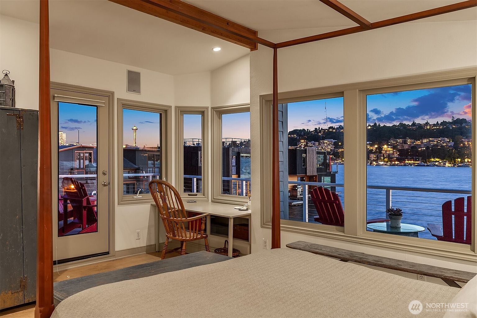This is a bedroom with a four-poster bed, featuring a view of the water and city skyline through multiple windows. The room includes a small desk area and a neutral color palette, creating a serene and inviting atmosphere. The overall impression is one of luxury and tranquility, with a focus on the stunning waterfront view.