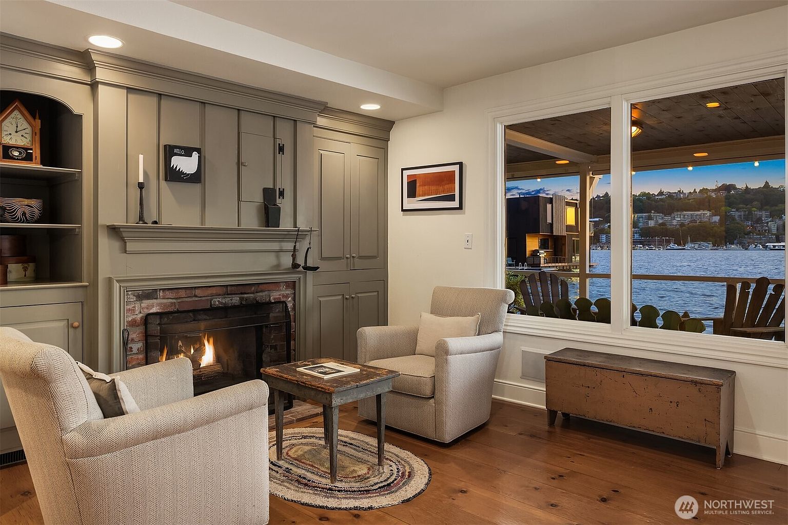 This inviting living room features a cozy fireplace with a brick surround and a painted mantel, flanked by built-in shelving and cabinetry. Two comfortable armchairs are arranged around a small rustic table on a braided rug, creating a warm and intimate seating area. A large window offers a picturesque view of the water, enhancing the room's serene atmosphere.