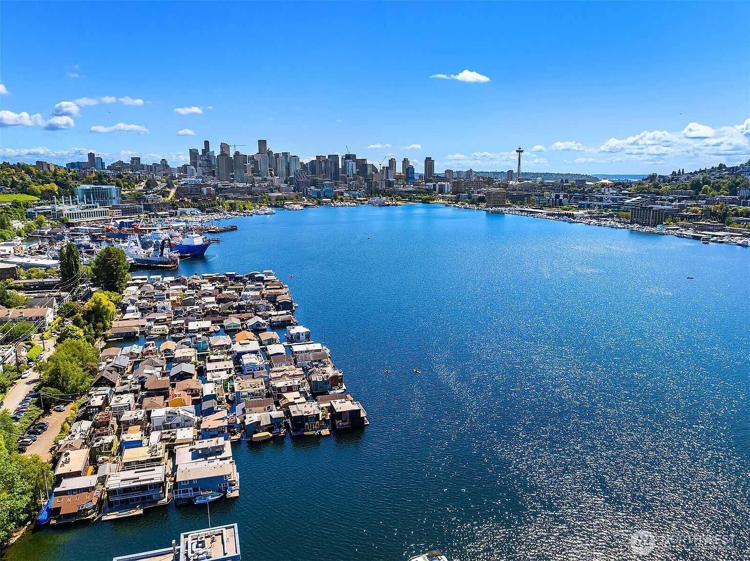 This aerial view showcases a unique floating home community on a vibrant blue lake, set against the backdrop of a bustling city skyline. The cluster of houseboats creates a charming and unconventional neighborhood, offering a distinctive waterfront living experience. The clear blue sky and sparkling water enhance the picturesque setting, highlighting the appeal of this urban oasis.
