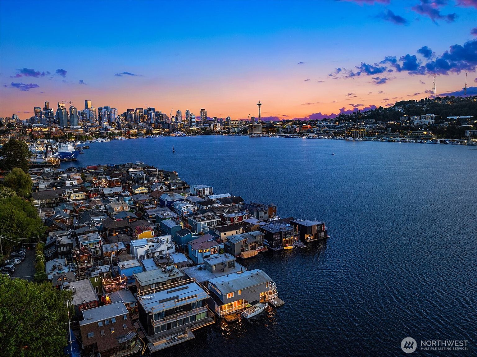 This aerial view showcases a unique floating home community on a serene lake, with the Seattle skyline and Space Needle providing a stunning backdrop at dusk. The homes are clustered together, creating a vibrant and picturesque neighborhood on the water, while the city lights reflect on the calm surface of the lake, enhancing the overall appeal and highlighting the desirable location.