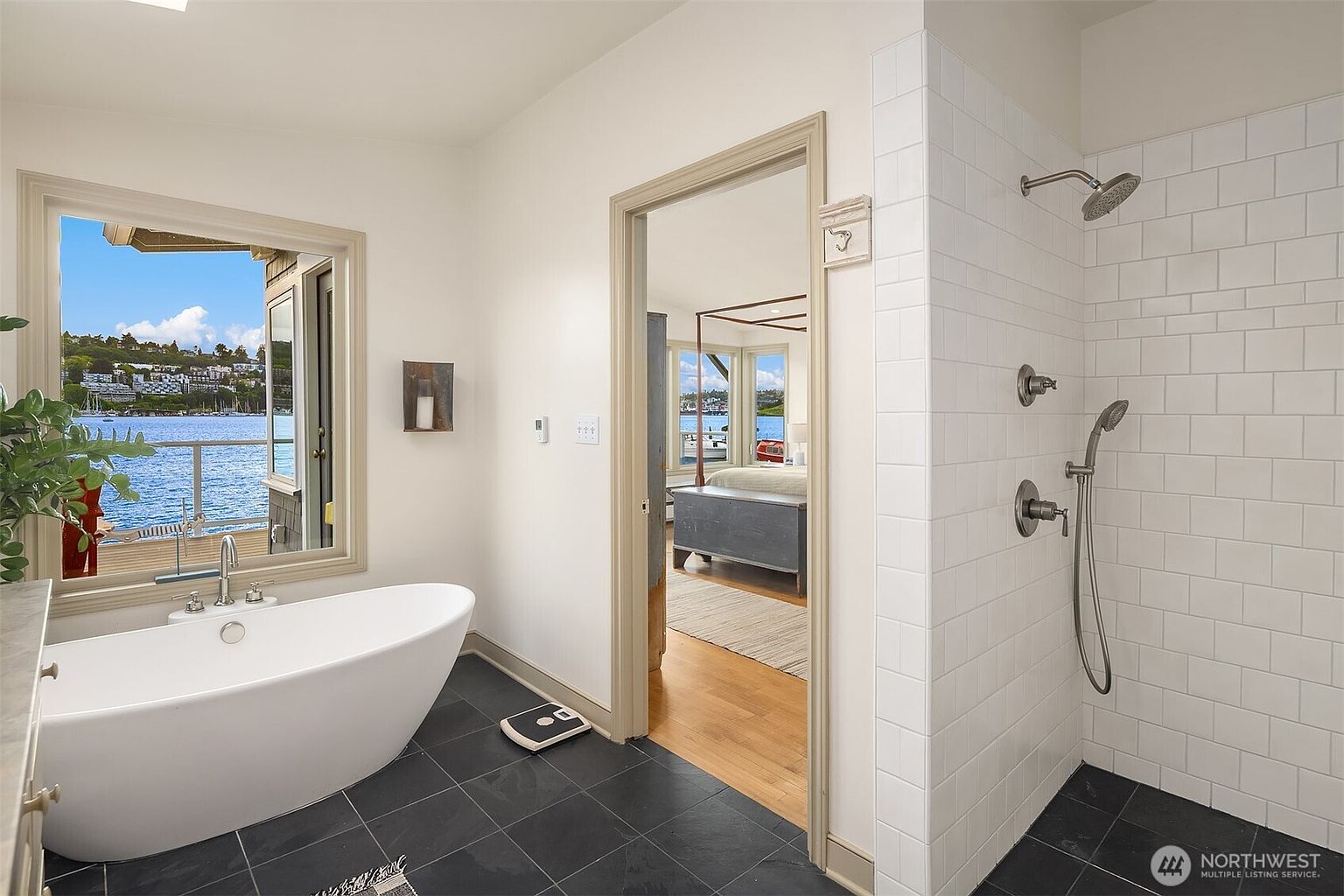 This is a bright and airy primary bathroom featuring a freestanding white bathtub positioned near a window with a waterfront view. The bathroom has dark tile flooring, a walk-in shower with white subway tiles, and a doorway leading to the primary bedroom. The overall design is modern and luxurious, emphasizing natural light and serene views.