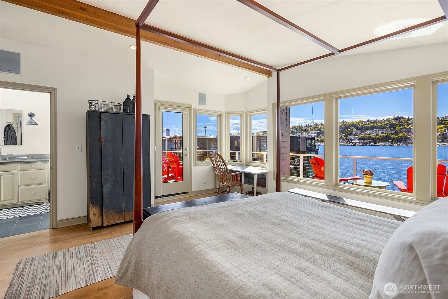 This primary bedroom features a four-poster bed with a neutral-toned comforter, hardwood floors, and large windows offering a waterfront view. The room includes a small desk area and an antique-style wardrobe. The overall impression is bright, airy, and luxurious, with a focus on the scenic view.