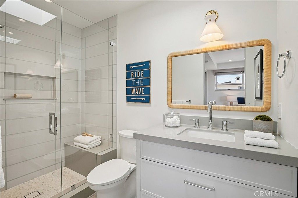 This bright and modern bathroom features a walk-in shower with a glass enclosure and light-colored wall tiles. A vanity with a stone countertop and a rectangular sink sits beneath a decorative rattan-framed mirror and a single pendant light. A wall-mounted sign adds a playful coastal touch to the clean, neutral space.