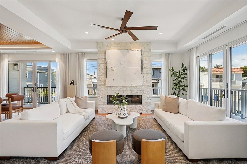 This bright and airy living room features a symmetrical layout with two white sofas facing each other across a central, organic-shaped coffee table. A prominent stone-clad chimney breast serves as the focal point, housing a linear fireplace and a large piece of abstract art. The space is framed by large glass doors on either side that provide access to outdoor balconies, while a modern ceiling fan hangs from the center of the room.