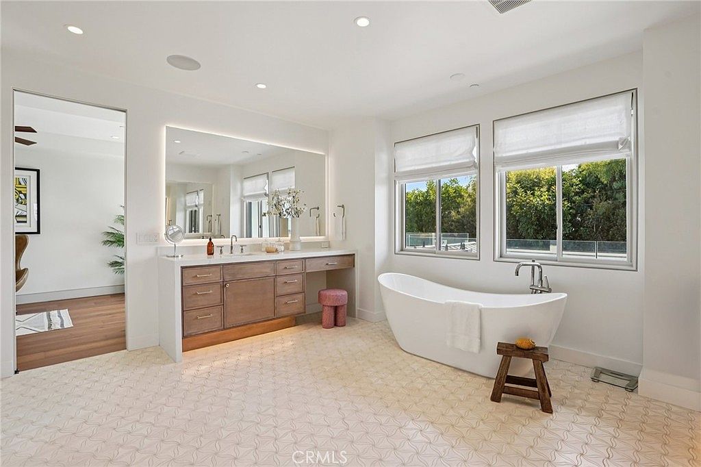 This bright and airy primary bathroom features a freestanding soaking tub positioned beneath large windows that offer a view of the outdoors. A long vanity with light wood cabinetry and a large, wide mirror provides ample space, complemented by a small pink stool. The room is finished with patterned tile flooring, creating a clean, modern, and luxurious aesthetic.