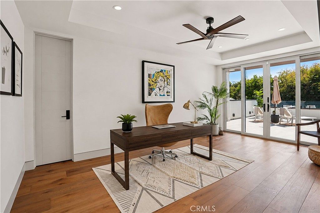 This bright and modern home office features warm wood flooring and a geometric patterned area rug beneath a dark wood desk. A tan leather chair sits at the desk, which is accented by a small desk lamp and a potted plant, while a piece of colorful pop art hangs on the white wall. Large glass doors open onto a sunny outdoor balcony, creating a seamless transition between the indoor workspace and the exterior living area.
