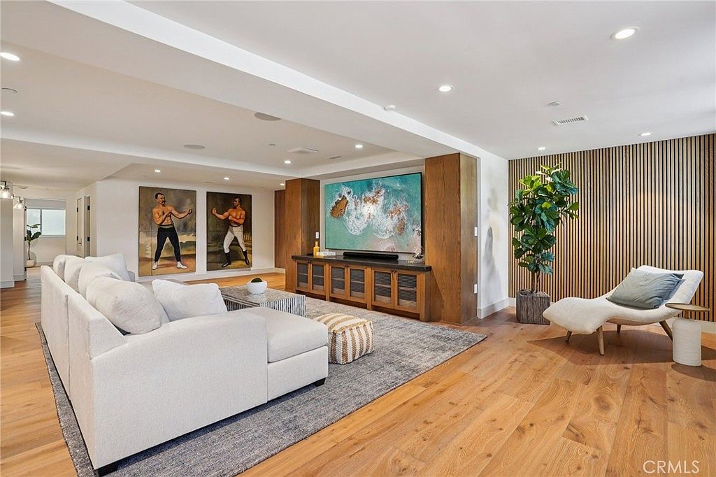 This spacious living room features a large, light-colored sectional sofa arranged on a textured area rug, creating a comfortable seating area. A media console sits beneath a large television, flanked by wood-paneled walls and two framed portraits of boxers. The room is accented by a modern lounge chair, a potted plant, and warm recessed lighting, offering a sophisticated and inviting atmosphere.
