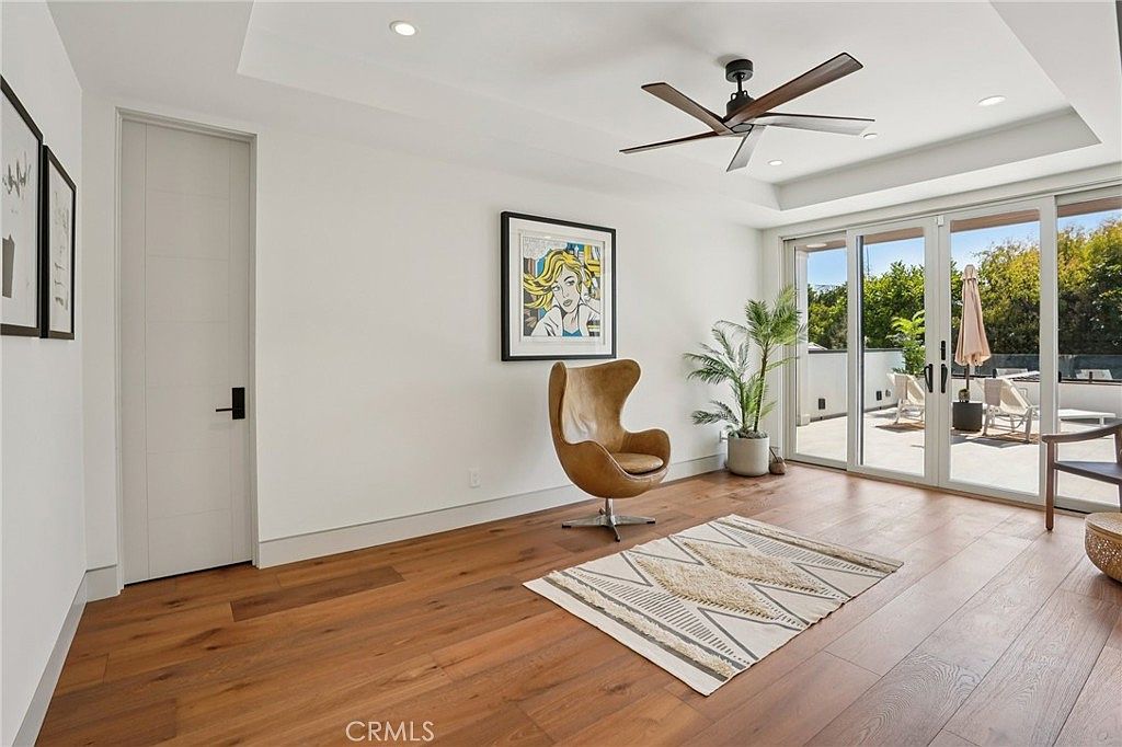 This bright and modern living space features warm-toned wood flooring and a neutral color palette. A stylish leather accent chair sits in the center of the room, complemented by a patterned area rug and a piece of pop-art on the wall. Large glass doors open onto an outdoor balcony, allowing for an abundance of natural light and a seamless transition to the exterior.