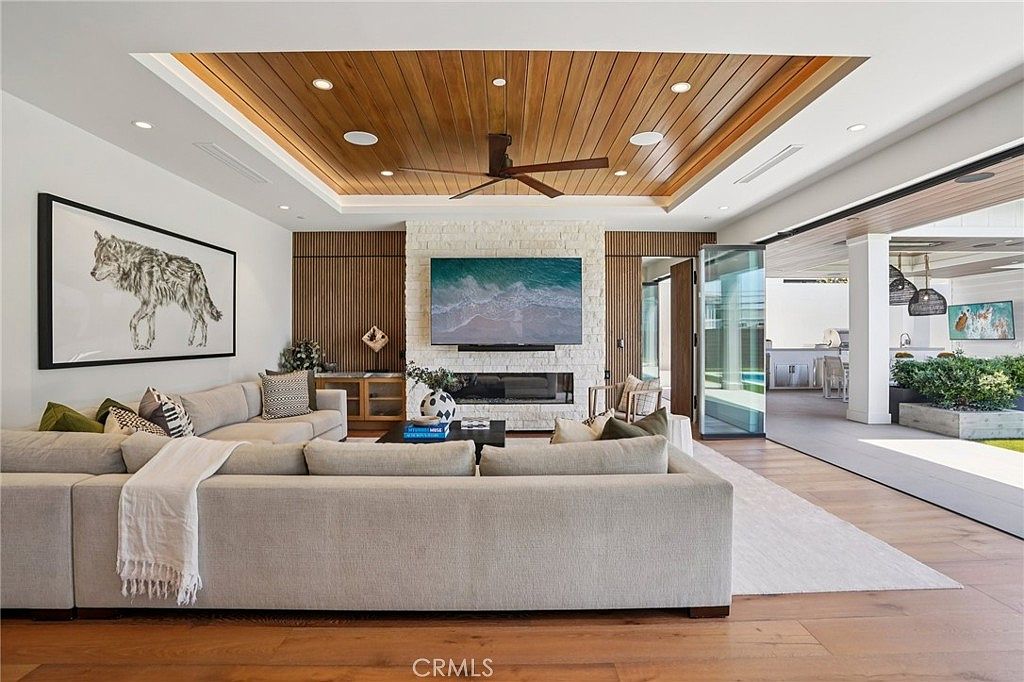 This spacious, open-concept living room features a large, light-toned sectional sofa facing a central stone-clad chimney breast with an integrated television and fireplace. The room is highlighted by a recessed wood-paneled ceiling with a central fan and recessed lighting, creating a warm, modern aesthetic. Large sliding glass doors open to an expansive covered patio, seamlessly blending indoor and outdoor living spaces.