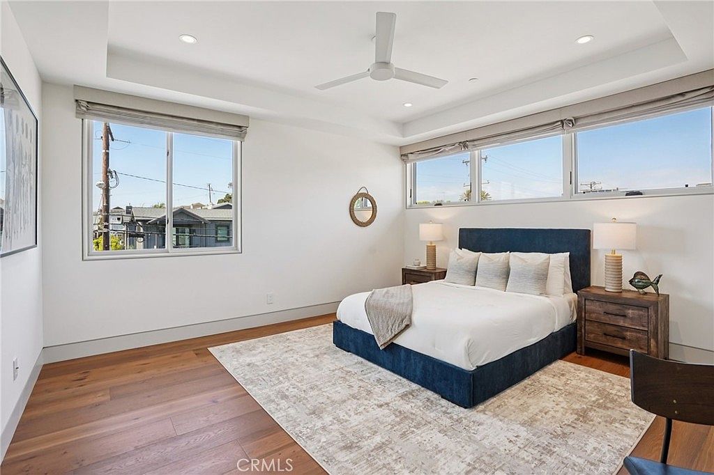 This bright and airy primary bedroom features light-toned wood flooring and a large, neutral-patterned area rug centered under a blue upholstered bed frame. The room is illuminated by recessed lighting and a ceiling fan, with large windows providing ample natural light and views of the surrounding neighborhood. The minimalist aesthetic is complemented by two matching nightstands with table lamps, creating a clean and tranquil atmosphere.