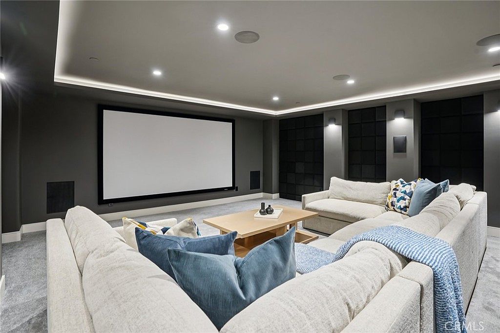 This dedicated home theater room features a large projection screen mounted on a dark accent wall, complemented by recessed cove lighting and integrated ceiling speakers. A spacious, light-colored sectional sofa with blue accent pillows provides ample seating, centered around a light wood coffee table. The room is designed for an immersive cinematic experience with a neutral color palette and plush carpeting.
