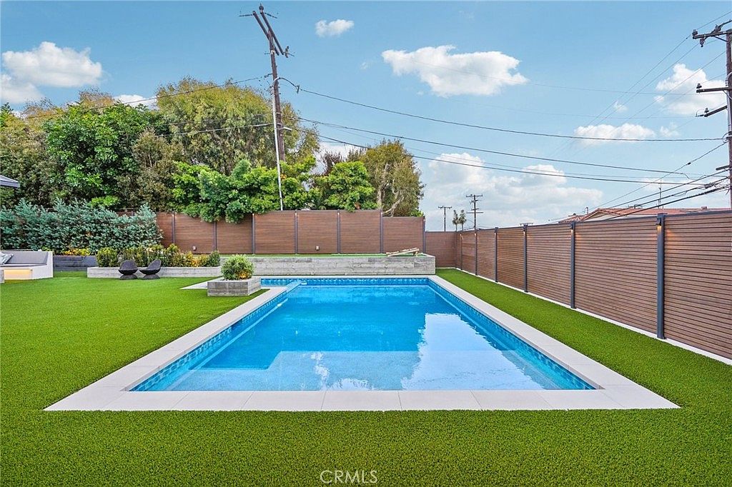 This backyard features a rectangular swimming pool as the central focal point, surrounded by a vibrant green artificial turf lawn. The area is enclosed by modern horizontal wood-slat fencing, providing a private and contemporary outdoor living space. The perspective is a wide-angle shot from the lawn, capturing the clean lines of the pool and the surrounding landscaping under a bright, partly cloudy sky.