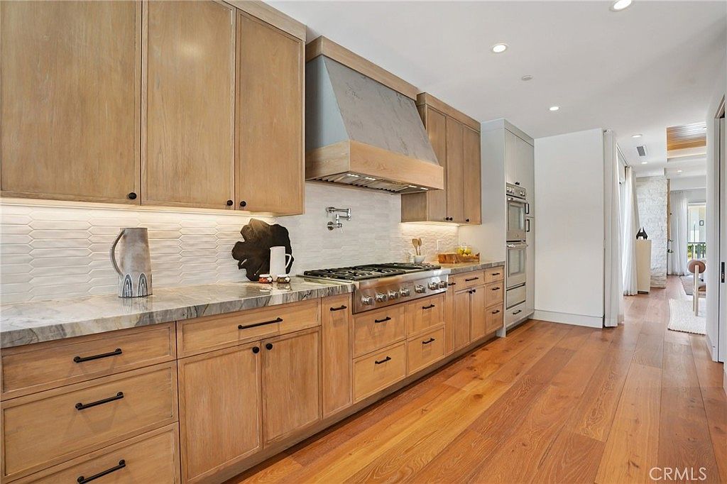 This modern kitchen features light wood cabinetry paired with a stone countertop and a textured tile backsplash. A professional-grade gas range is centered beneath a custom-built range hood, while warm wood flooring extends throughout the space. The perspective captures the length of the kitchen, highlighting the clean lines and the transition into an adjacent hallway.