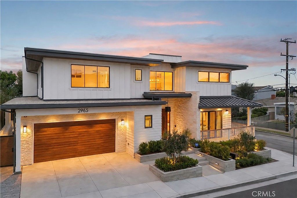 This modern two-story residence features a striking blend of white vertical siding and textured stone accents, creating a sophisticated curb appeal. The front facade includes a spacious garage with a warm-toned wood door, a welcoming entryway with a covered porch, and neatly landscaped tiered planters. Captured at dusk, the warm exterior lighting highlights the architectural lines and inviting atmosphere of the home.