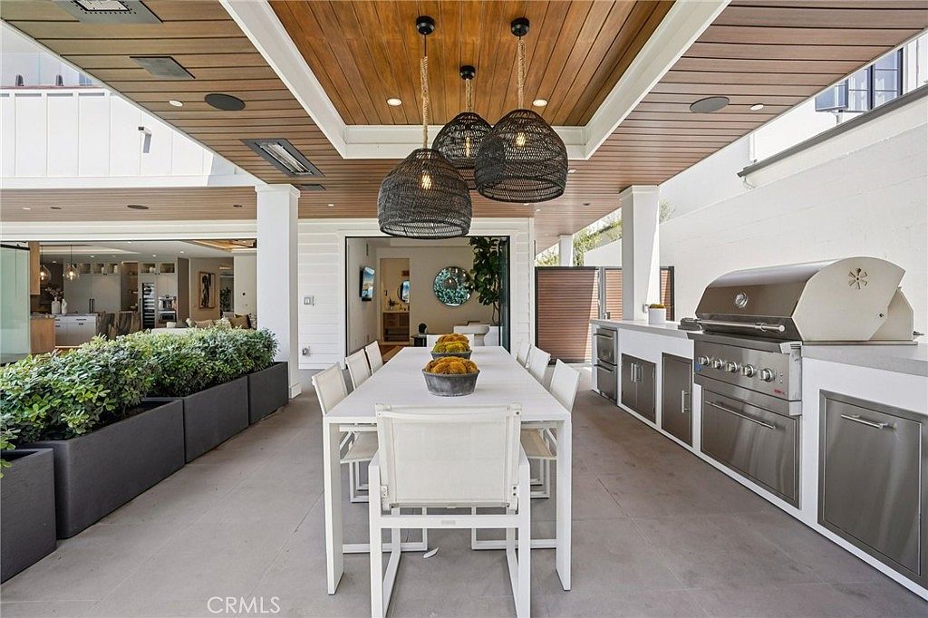 This sophisticated covered outdoor dining area features a long white table surrounded by matching chairs, positioned beneath a warm wood-paneled ceiling with three large woven pendant lights. To the right, a built-in outdoor kitchen includes a stainless steel grill and ample counter space, while a row of lush green planters lines the left side. The space seamlessly transitions to the interior living area through wide-opening glass doors, creating an expansive and airy atmosphere perfect for entertaining.