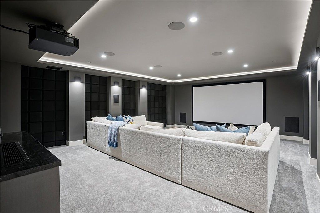 This modern home theater features a large projection screen mounted on a dark accent wall, complemented by a spacious, light-colored sectional sofa that provides ample seating. The room is designed with recessed ceiling lighting and dark, textured wall panels, creating a sophisticated and immersive cinematic environment. A projector is mounted on the ceiling, and a dark countertop area is visible in the foreground, adding to the functional layout of this dedicated entertainment space.