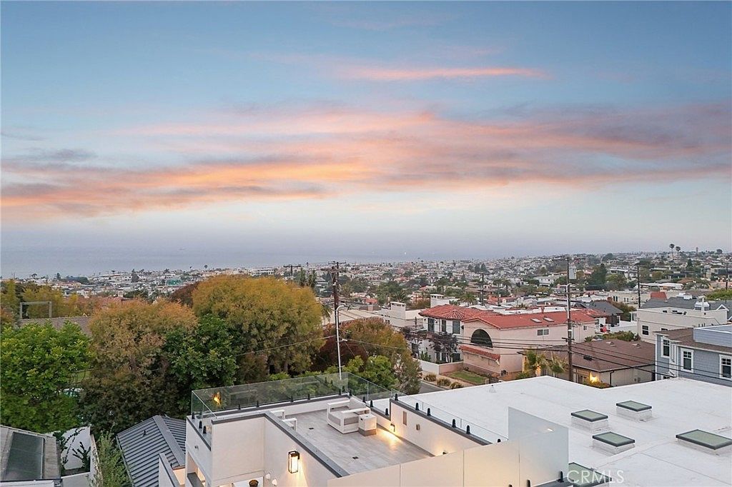 This elevated perspective captures a sweeping panoramic view of a coastal city at dusk, with the horizon meeting a soft, pastel-colored sky. In the foreground, a modern rooftop deck features glass railings and outdoor seating, overlooking a dense residential neighborhood and the distant ocean. The scene emphasizes the property's prime location and expansive outdoor living space.