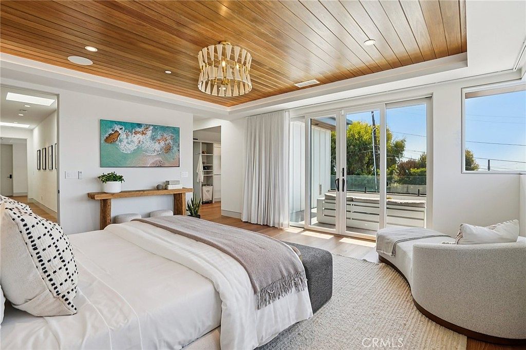 This primary bedroom features a striking wood-paneled ceiling with a decorative chandelier and large glass doors that open to a private balcony. The room is furnished with a plush bed, a curved chaise lounge, and a rustic wood console table beneath a coastal-themed artwork. The neutral color palette and natural textures create a serene, high-end atmosphere with a seamless transition to the outdoor space.