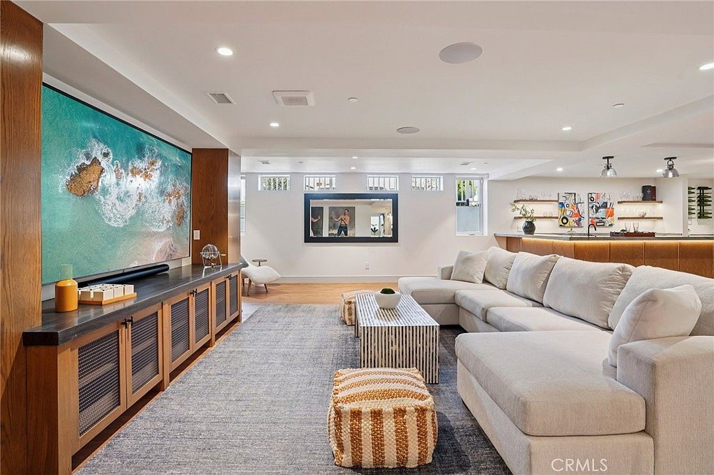 This spacious, open-concept living area features a large, light-colored sectional sofa arranged around a patterned coffee table and a textured rug. A massive wall-mounted television is set into a custom wood media console, while the background reveals a bar area with open shelving and recessed lighting. The room is bright and airy, with a neutral color palette and a contemporary design that flows seamlessly into the adjacent kitchen space.