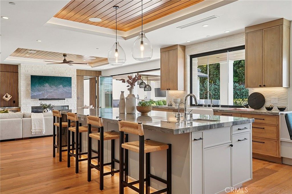 This bright, open-concept kitchen features a large central island with a stone countertop and bar seating, illuminated by two glass pendant lights. The space showcases light wood cabinetry, a white farmhouse-style sink, and a seamless transition to the adjacent living area, which includes a large television mounted above a stone-clad chimney breast. The room is characterized by warm wood flooring and a wood-paneled ceiling, creating an inviting and airy atmosphere.