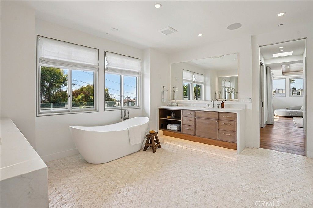 This bright and airy primary bathroom features a freestanding soaking tub positioned beneath large windows that allow for ample natural light. A long vanity with light wood cabinetry and a stone countertop provides generous storage, complemented by a large wall-mounted mirror. The space is finished with intricate patterned floor tiles and leads into an adjacent room with hardwood flooring, creating a cohesive and luxurious aesthetic.