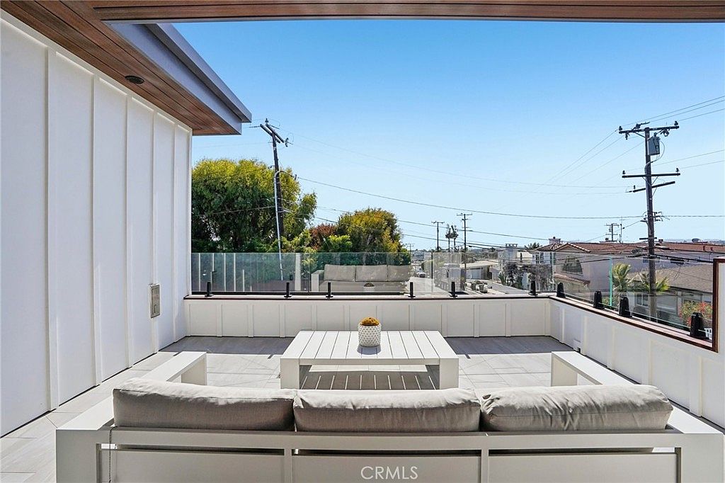 This modern rooftop deck features a clean, minimalist aesthetic with light-toned tile flooring and a white modular sofa arrangement surrounding a matching square coffee table. A glass railing provides an unobstructed view of the surrounding neighborhood and utility lines, while a partial roof overhang offers shade. The space is designed for outdoor relaxation and entertaining, capturing a bright, airy atmosphere.
