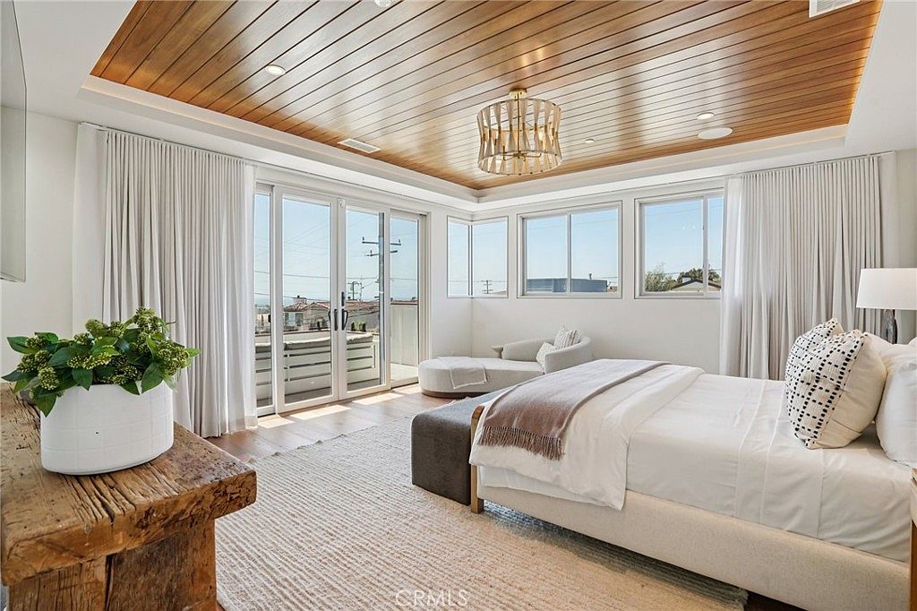 This spacious primary bedroom features a striking wood-paneled ceiling and a neutral color palette that creates a serene atmosphere. The room is anchored by a large bed with a textured throw, complemented by a rustic wood console table and a cozy lounge area near the windows. Large glass doors open to a private balcony, allowing for abundant natural light and a seamless connection to the outdoors.