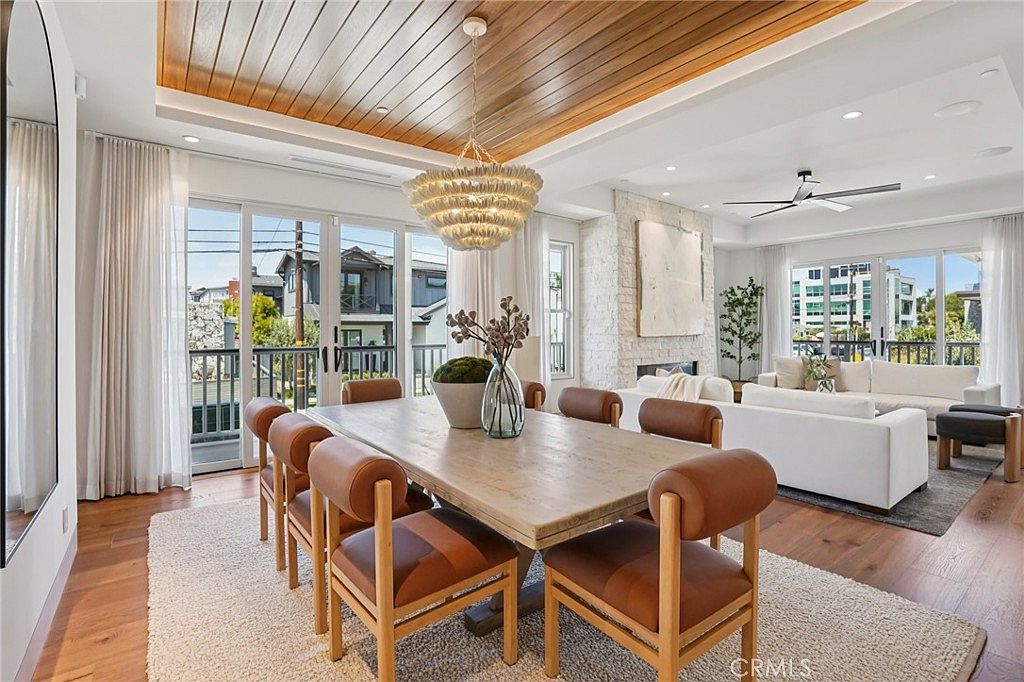 This open-concept dining area features a light wood table surrounded by chairs with brown leather upholstery, set atop a textured area rug. Above, a unique, textured chandelier hangs from a warm wood-paneled ceiling, while the adjacent living space showcases a stone-clad chimney breast and large glass doors leading to a balcony. The room is bathed in natural light, creating an airy and sophisticated atmosphere that seamlessly connects the dining and living zones.