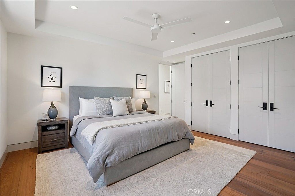 This spacious primary bedroom features a neutral color palette with a large upholstered bed as the central focal point. The room is accented by two matching bedside tables with lamps, framed wall art, and a ceiling fan, all set against light-toned walls and warm wood flooring. A large area rug anchors the space, while double-door closets provide ample storage, creating a clean and serene atmosphere.