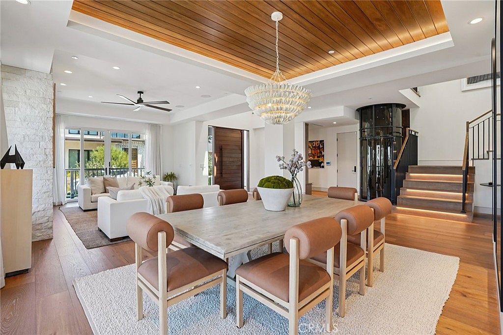 This open-concept dining area features a light wood table surrounded by six chairs with tan leather upholstery, set atop a textured area rug. A striking tiered chandelier hangs from a warm wood-paneled ceiling, while the adjacent living space includes white sofas and large windows that provide ample natural light. The room also showcases a modern glass-enclosed elevator and a staircase with illuminated steps, creating a sophisticated and airy atmosphere.
