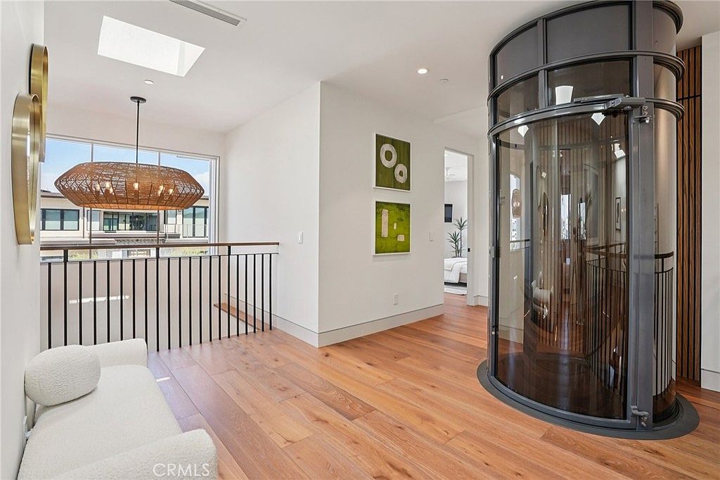 This modern landing area features light wood flooring and a striking cylindrical glass elevator that serves as a central architectural focal point. To the left, a woven pendant light hangs above a white sofa, while a black metal railing overlooks the level below. The space is bright and airy, with clean white walls and two framed green art pieces adding a touch of color to the hallway.