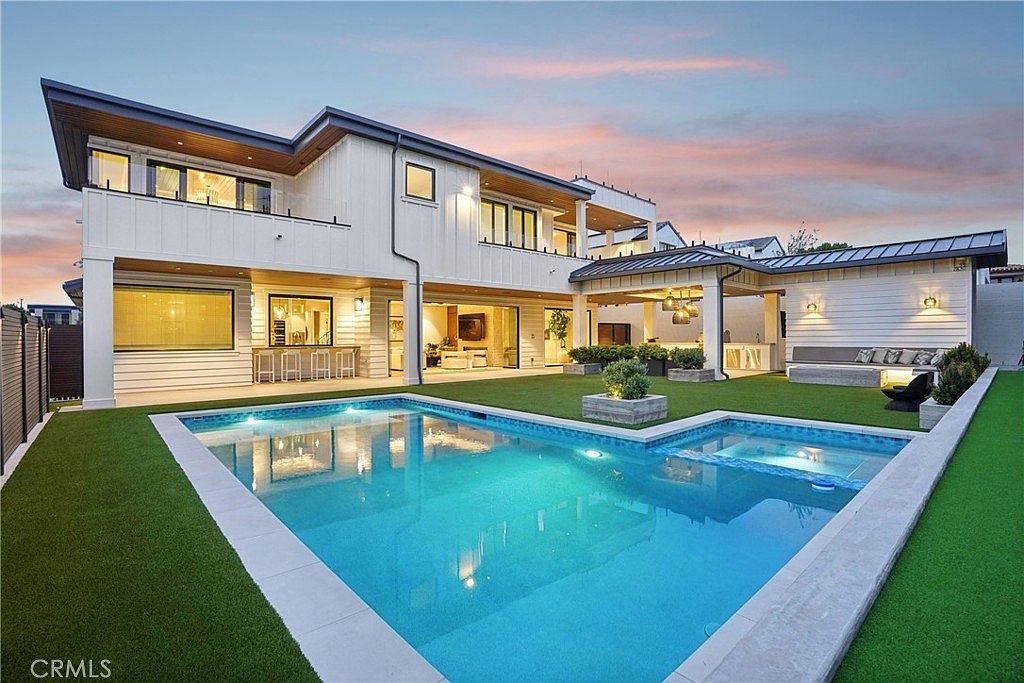 This modern luxury residence features a striking rear exterior with white vertical siding and expansive glass doors that seamlessly connect the indoor living space to the backyard. The outdoor area is highlighted by a pristine rectangular swimming pool with an integrated spa, surrounded by manicured artificial turf and a covered patio lounge. The scene is captured at dusk, with warm ambient lighting illuminating the home's architectural lines and the inviting outdoor entertainment zones.