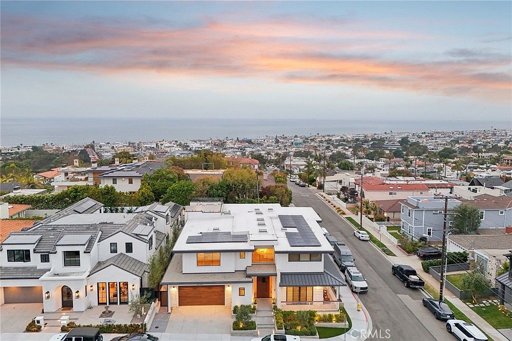 This high-angle aerial shot captures a contemporary two-story residence situated in a coastal neighborhood, showcasing its modern architectural design and solar panel-equipped roof. The property features a warm-toned wood garage door, a welcoming front porch, and manicured landscaping, all set against a backdrop of a sprawling suburban landscape meeting the ocean horizon at sunset. The perspective emphasizes the home's prominent position within the streetscape and its proximity to the scenic coastline.