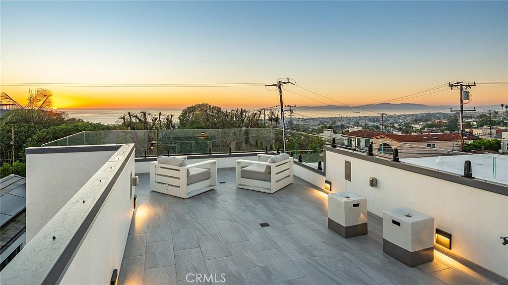 This rooftop deck offers a panoramic view of the ocean and surrounding coastal neighborhood at sunset. The space is furnished with two white lounge chairs and two square side tables, set against a modern glass railing. The deck is finished with light-toned, large-format tiles and features integrated low-level lighting for an inviting evening atmosphere.
