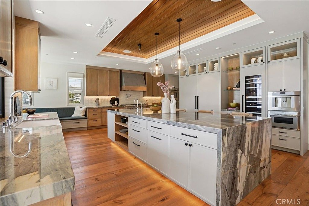 This expansive, high-end kitchen features a large central island with a dramatic stone countertop and waterfall edge, complemented by light wood cabinetry and white upper cabinets. A striking wood-paneled ceiling detail adds warmth to the space, while two glass pendant lights hang above the island. The room is finished with warm-toned wood flooring and integrated high-end appliances, creating a sophisticated and cohesive culinary environment.