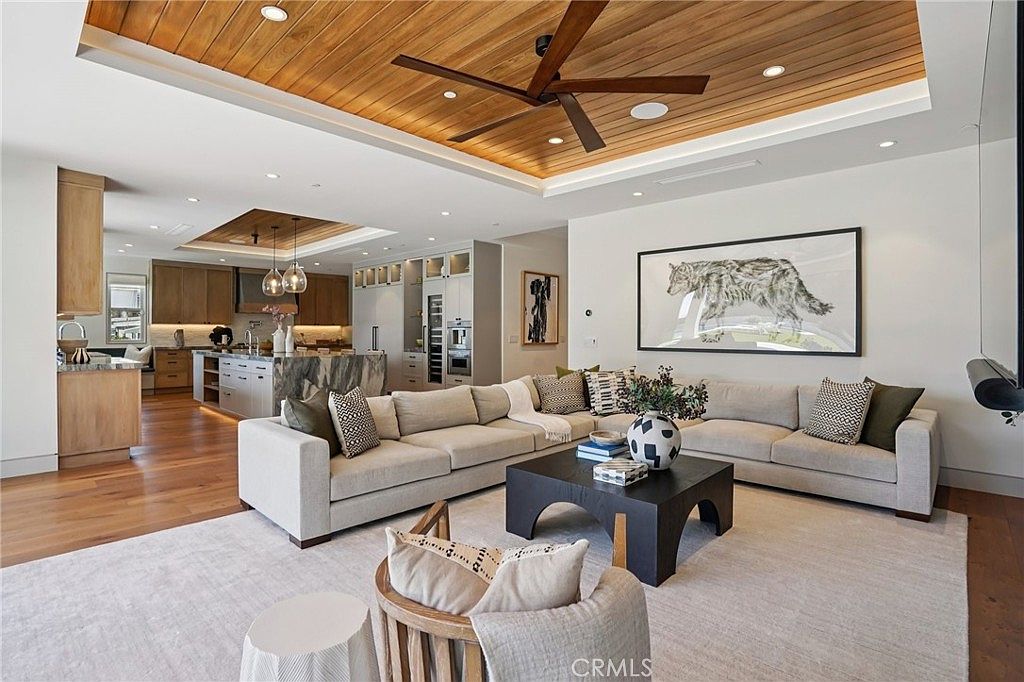 This open-concept living area features a large, neutral-toned sectional sofa arranged around a dark, geometric coffee table. The space is defined by a coffered ceiling with warm wood paneling and a modern ceiling fan, creating a sophisticated and inviting atmosphere. In the background, a spacious kitchen with a large stone-topped island and light wood cabinetry is visible, emphasizing the seamless flow between living and dining areas.