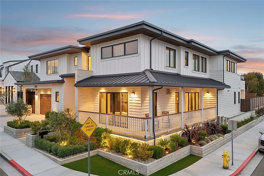 This modern two-story residence features a striking blend of white vertical siding and horizontal lap siding, accented by a dark metal roof and warm exterior lighting. The home is situated on a corner lot, showcasing a wrap-around porch with a white railing and a neatly landscaped garden area. The perspective is a low-angle, wide shot that captures the architectural details and the inviting glow of the interior lights against the twilight sky.