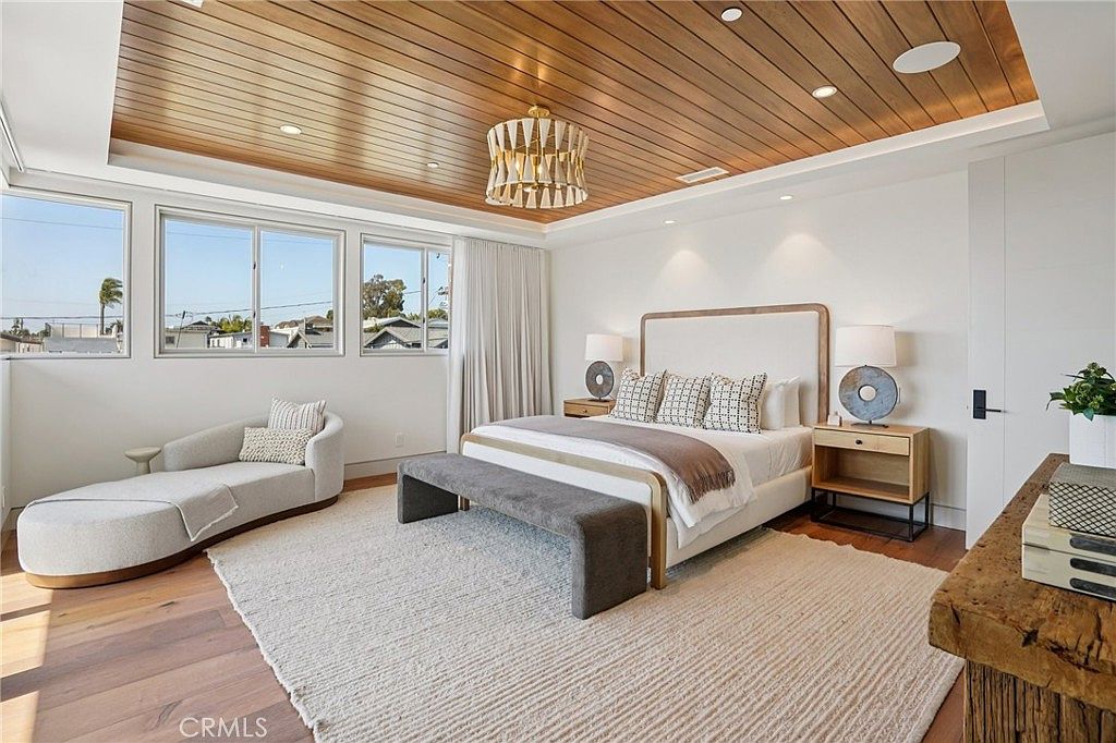 This spacious primary bedroom features a warm wood-paneled ceiling with a modern gold-toned chandelier and light wood flooring. The room is anchored by a large bed with a neutral upholstered headboard, flanked by two wooden nightstands, and includes a curved chaise lounge positioned near a series of windows. The overall aesthetic is bright and airy, with a soft, neutral color palette that creates a serene and inviting atmosphere.