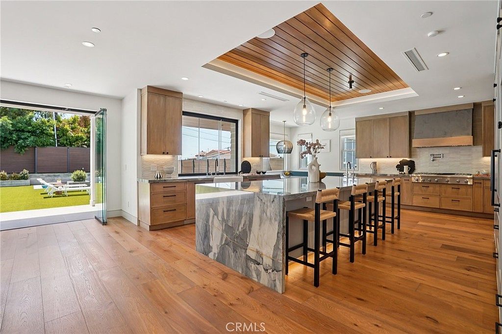 This expansive, modern kitchen features a large central island with a waterfall-edge stone countertop and seating for four. The space is defined by light wood cabinetry, a recessed wood-paneled ceiling detail with pendant lighting, and wide-plank wood flooring. Large glass doors open directly to an outdoor lawn area, creating a seamless transition between the indoor cooking space and the exterior yard.