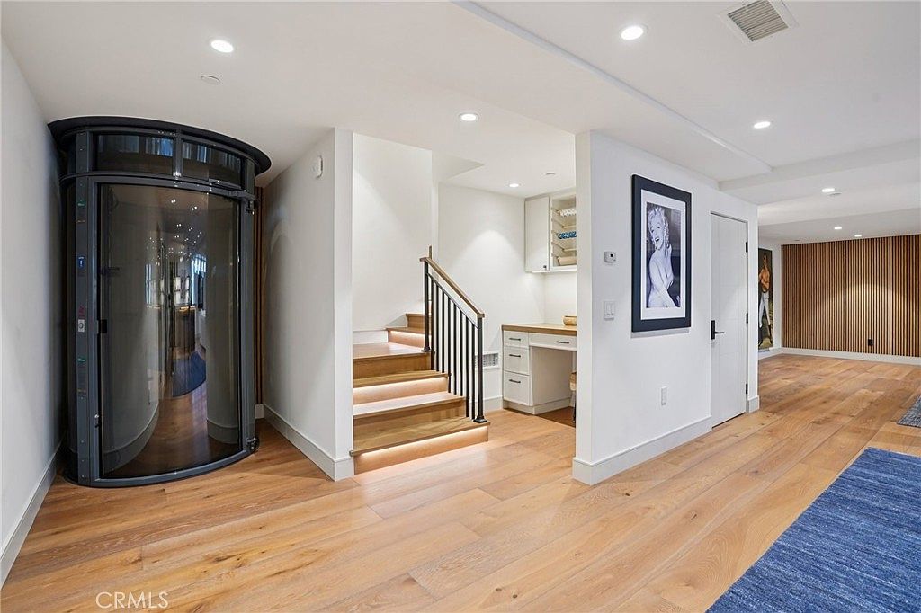 This interior view showcases a modern hallway featuring a cylindrical glass elevator on the left and a staircase with light wood treads and a black metal railing. To the right, a built-in desk area is tucked under the stairs, while a hallway leads toward the back of the home with wood-paneled walls. The space is bright and contemporary, characterized by clean lines, recessed lighting, and light-toned wood flooring throughout.