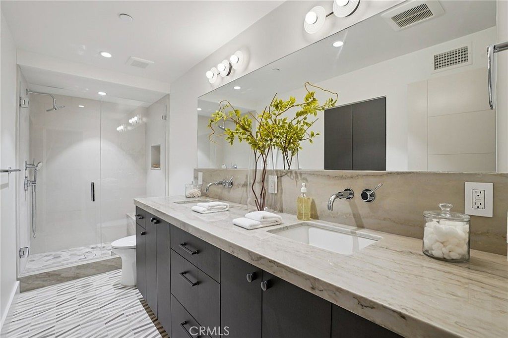 This modern primary bathroom features a dual-sink vanity with dark cabinetry and a light-toned stone countertop. A large, frameless glass-enclosed shower is positioned to the left, while a wide mirror spans the wall above the vanity, reflecting the bright, clean space. The room is finished with patterned tile flooring and contemporary lighting fixtures, creating a sleek and sophisticated atmosphere.