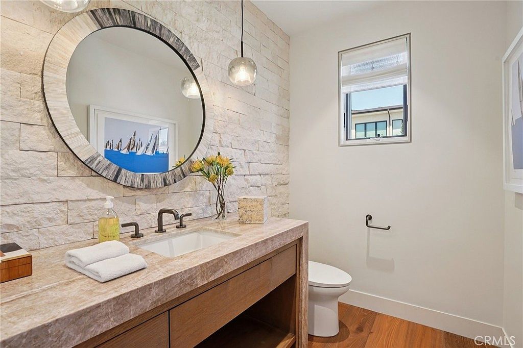 This elegant powder room features a textured stone accent wall behind a floating vanity with a stone countertop and an integrated sink. A large, circular mirror with a decorative frame hangs above the vanity, complemented by a modern pendant light and a small vase of flowers. The space is finished with warm wood flooring and a clean, neutral color palette, creating a sophisticated and inviting atmosphere.