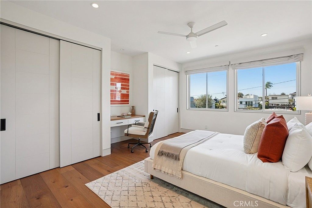 This bright and airy bedroom features light wood flooring, a neutral color palette, and a built-in desk area tucked into a recessed nook. A large bed with white linens and warm-toned accent pillows serves as the focal point, while two large windows allow for ample natural light and views of the neighborhood. The room is finished with a modern ceiling fan and clean, minimalist lines, creating a serene and functional living space.