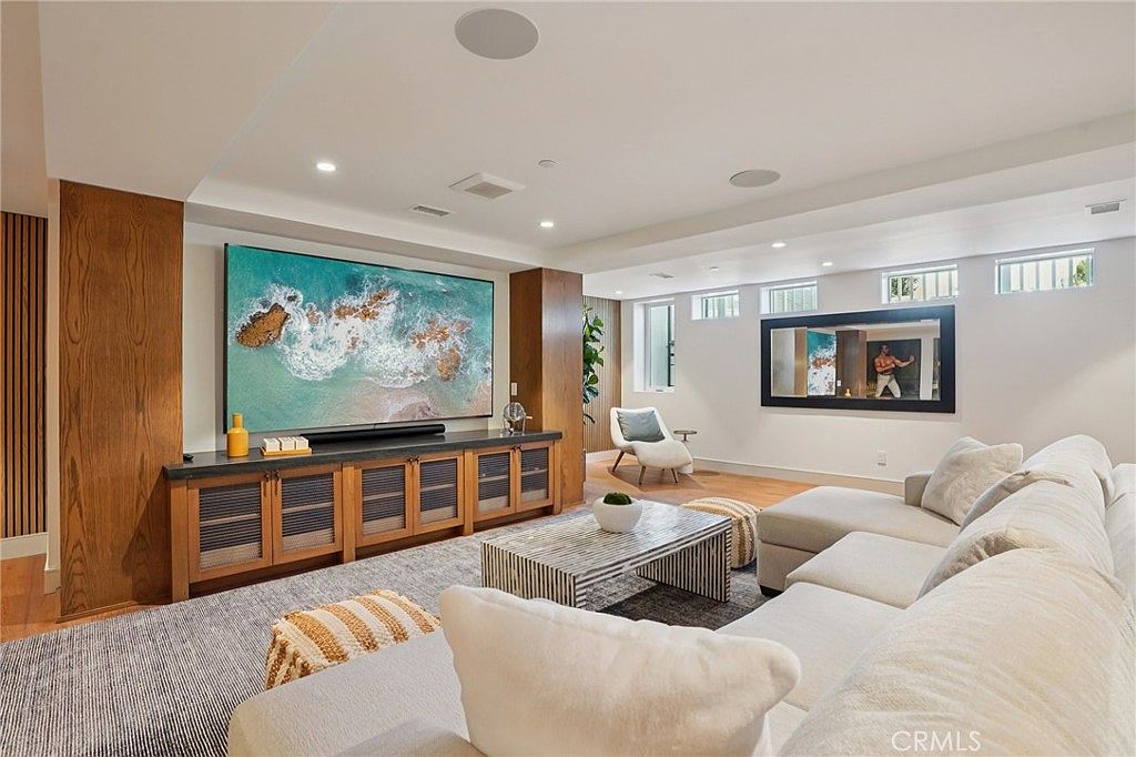 This spacious, modern living room features a large television mounted on a wood-paneled wall above a long, low-profile media console. A plush, light-colored sectional sofa faces the screen, centered around a patterned coffee table on a textured area rug. The room is brightened by recessed lighting and a series of high-set windows, creating an inviting and comfortable atmosphere for relaxation.