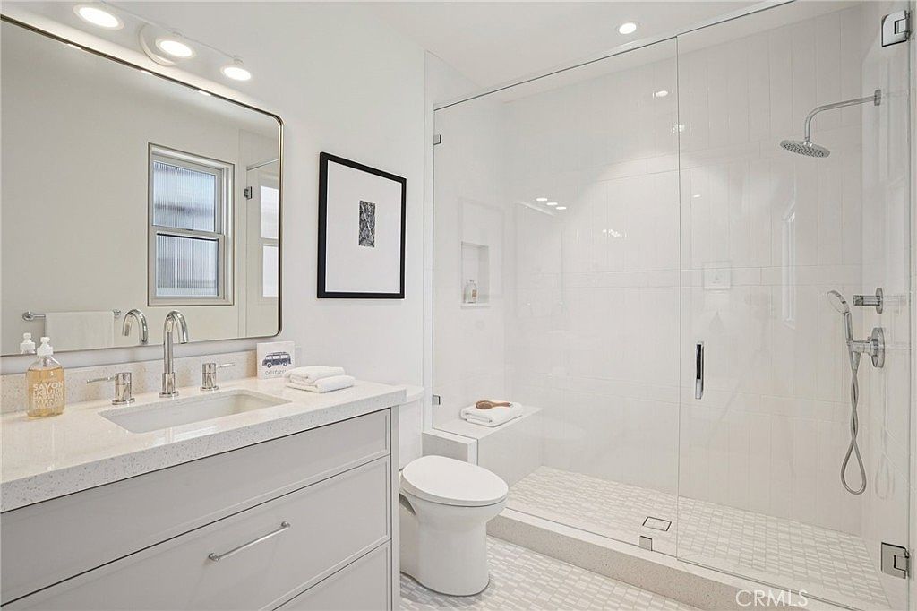 This bright, modern bathroom features a sleek vanity with a light-toned countertop and a rectangular mirror illuminated by a multi-bulb light fixture. To the right, a spacious walk-in shower is enclosed by a glass panel and includes a built-in bench, a rainfall showerhead, and a handheld sprayer. The room is finished with light-colored walls and patterned floor tiles, creating a clean and contemporary aesthetic.