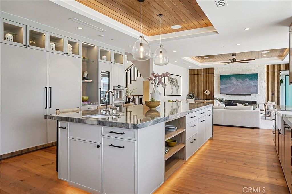 This expansive, open-concept kitchen features a large central island with a stone countertop and integrated seating, illuminated by two glass pendant lights. The space is defined by light-toned cabinetry, warm wood ceiling accents, and light wood flooring that extends into the adjacent living area. A stone-clad wall serves as a focal point in the background, housing a large television and creating a seamless transition between the culinary and relaxation zones.